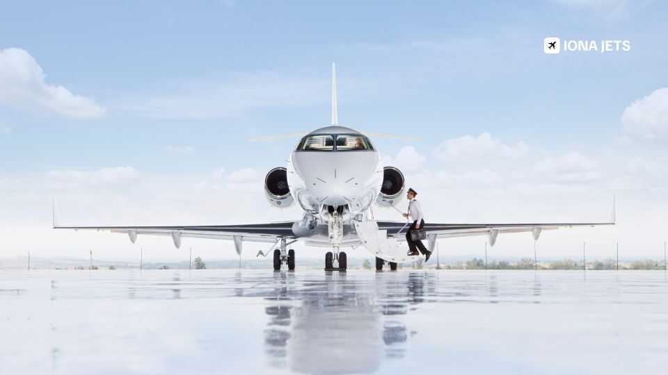 Front view of a white private jet parked on a wet runway under a bright sky, with a passenger boarding via the airstair and the IONA JETS logo in the top right corner.